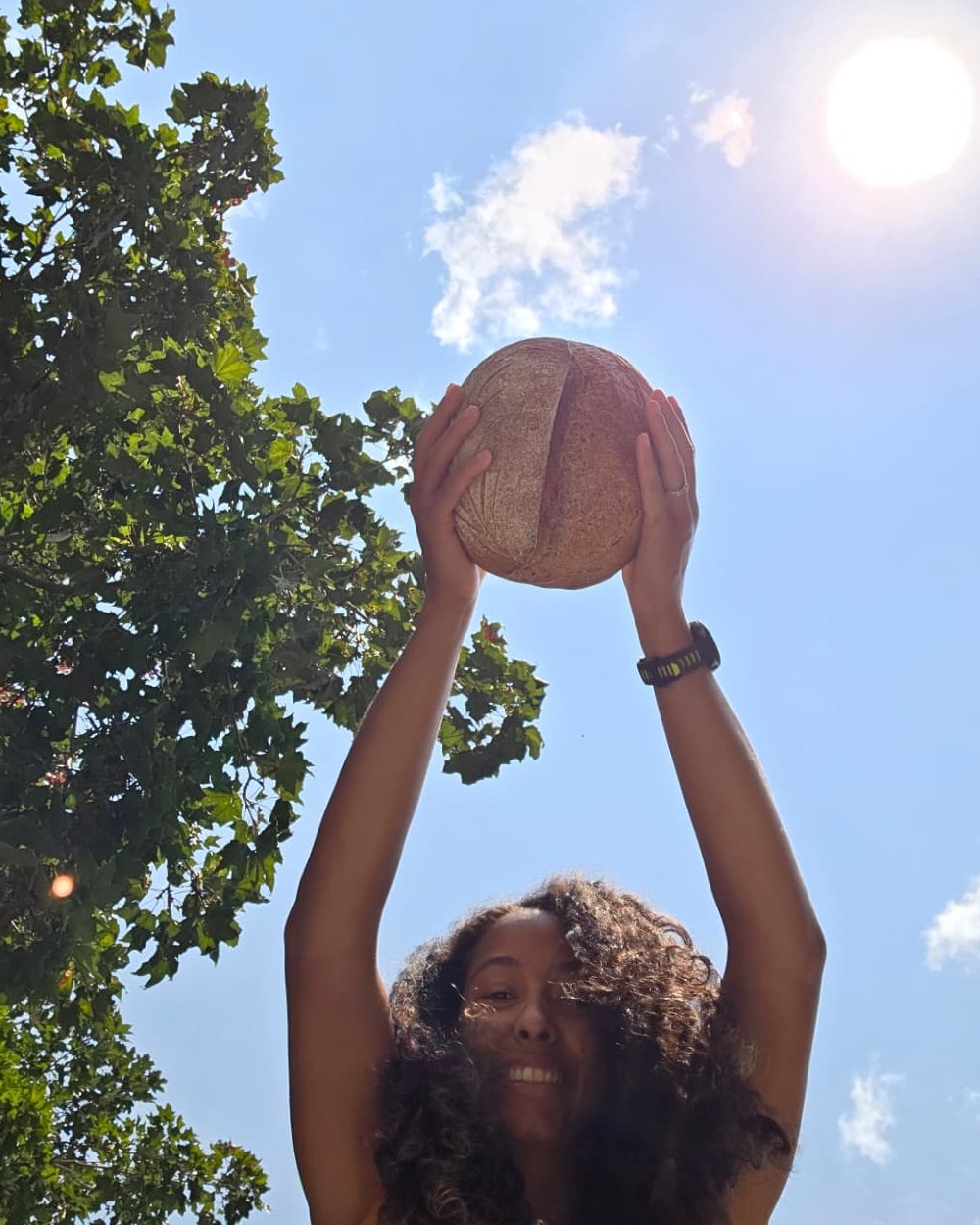 The baker behind Bread Actually holding a round loaf overhead beneath a blue sky